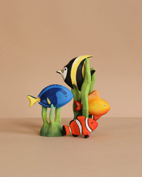 The Bumbu Handmade Wooden Tropical Fish Set features four colorful painted wooden fish—blue, yellow-black-white, orange, and yellow—standing among green plants on a beige background. Perfect addition to any Montessori toy collection.