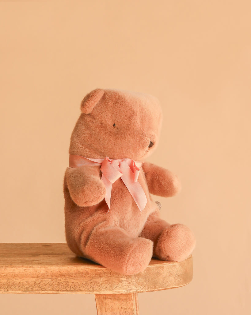 A Maileg Teddy Bear in Carry Cot sits on the edge of a wooden stool against a plain beige background, its soft brown plush and pink ribbon adding charm to the scene.