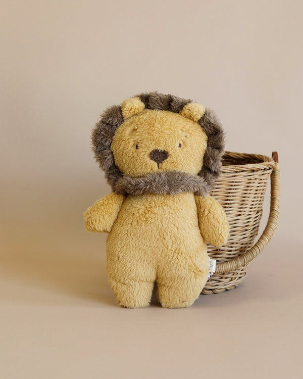 A soft, yellow Plush Lion Stuffed Animal with a fluffy mane made from recycled faux fur stands in front of a small, light brown woven basket on a beige background.