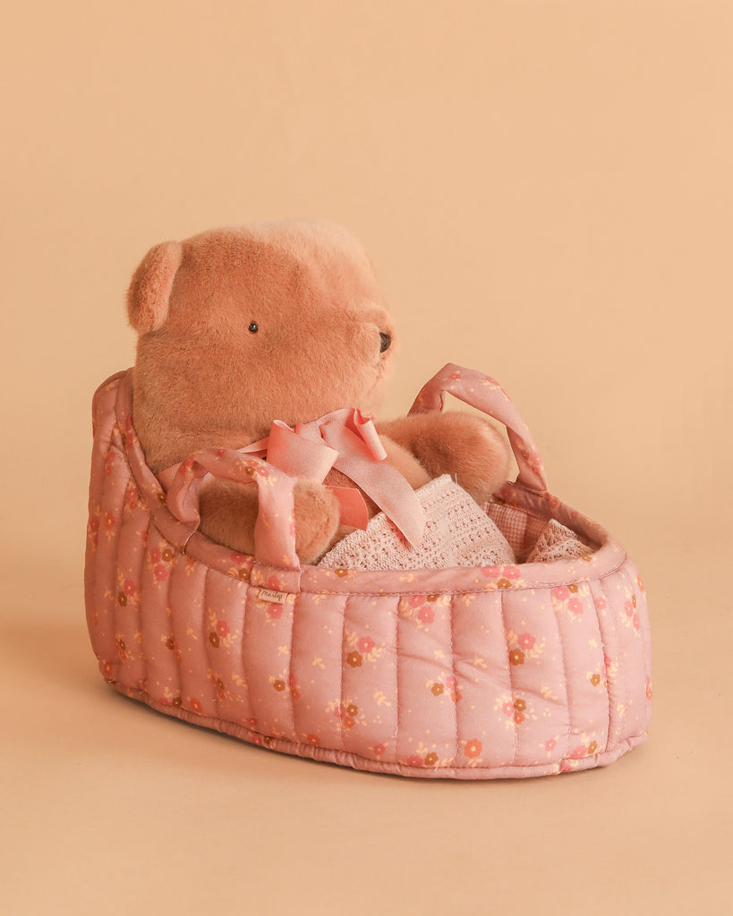 The Maileg Teddy Bear in Carry Cot, dressed in a white lace outfit, sits cozily in its pink floral-patterned cot against a peach backdrop.