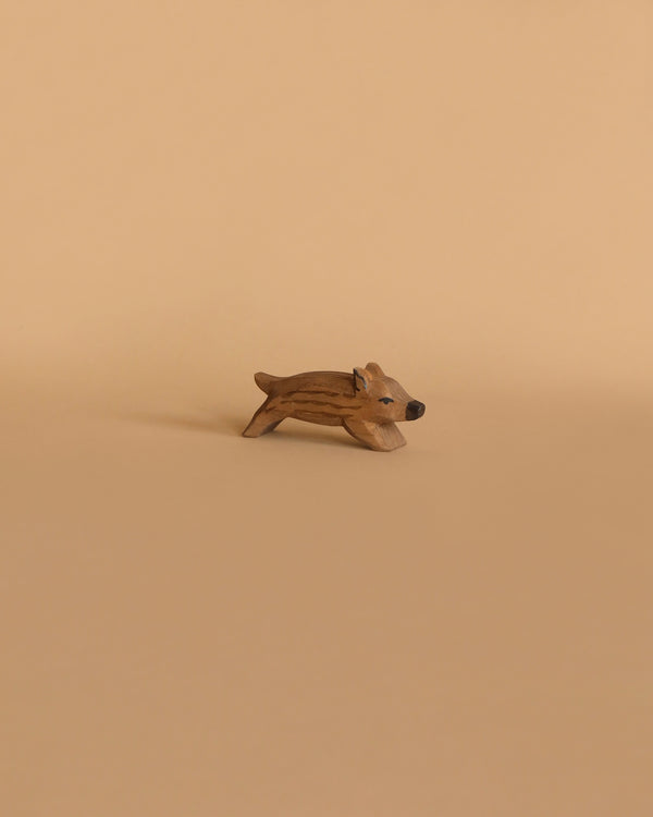 A small, light brown wooden figurine of a wild boar in a playful stance, handcrafted by Ostheimer, placed on a beige background.