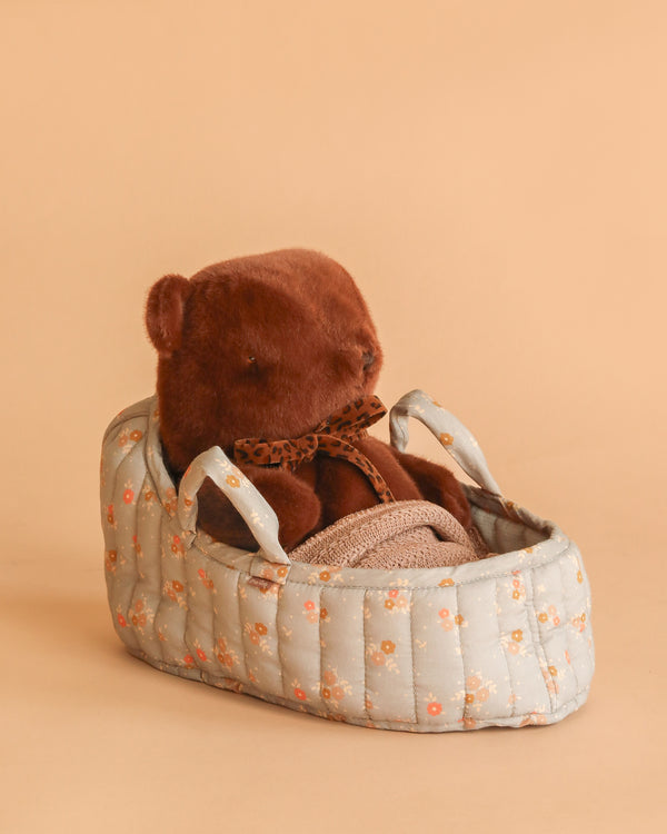 The Maileg Teddy Bear in Carry Cot sits with a leopard-print bow in a light blue, floral-patterned carrier against a solid light brown background.