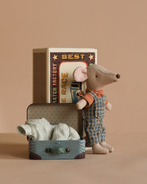 The Maileg Big Brother Mouse, dressed in checkered overalls and an orange-striped shirt, stands by a vintage-style suitcase filled with clothes. Nearby, a large matchbox displays "BEST." Set against a neutral beige background, it captures the charm of soft fabrics and childhood nostalgia.