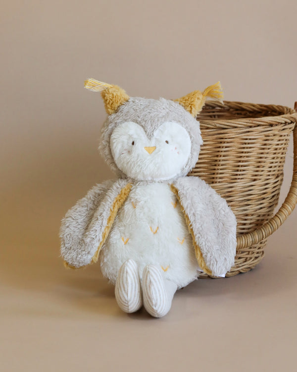 A Owl Stuffed Animal with a serene expression sits in front of a woven basket from Vietnam. The owl has soft grey and white fur, with little yellow details on its wings and ears.