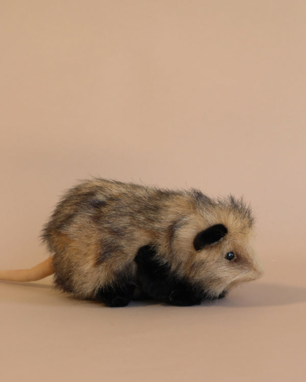 A realistic Opossum Stuffed Animal with mottled gray and black fur, standing on a plain beige background. This artisan crafted toy has a long, hairless pink tail, small ears, and.