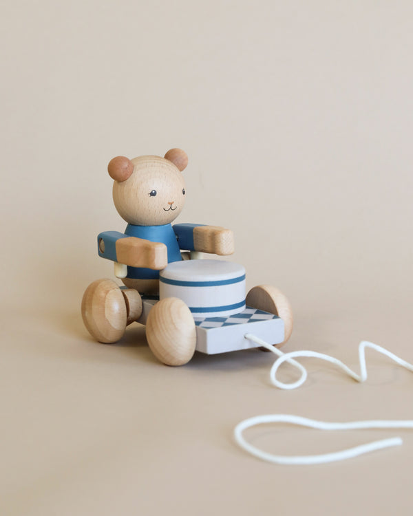 A Wooden Pull Toy - Drummer Bear made from FSC-certified wood, sitting on a blue and white toy car with wheels, against a soft beige background. The toy has a string attached to the front for pulling.