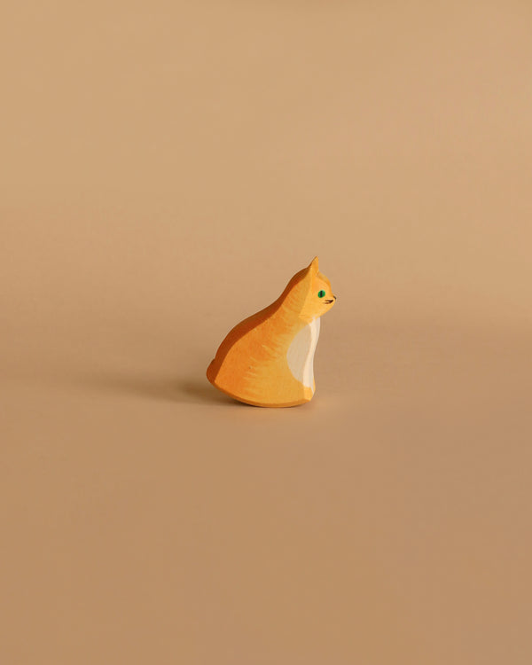 The Ostheimer Orange Cat, Sitting is a small, carved wooden figurine featuring an orange and white cat on a beige background. Reminiscent of traditional Ostheimer toys, this cat is depicted in a sitting position facing right. It has minimalistic features and a smooth finish, representing an excellent example of handcrafted wooden toys made from sustainably sourced materials.