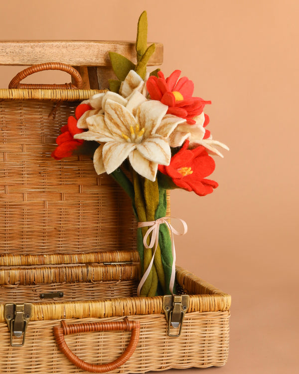 The Handcrafted Felt Flower Bouquet, featuring red and white wool-felt blooms tied with a pink ribbon, sits in an open wicker basket against a beige background.