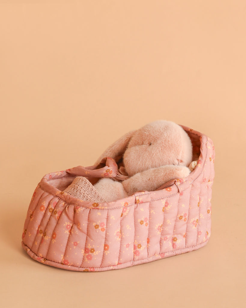 A Maileg Sleeping Bunny in Carry Cot rests in a quilted, pink floral-patterned bed against a soft peach background.