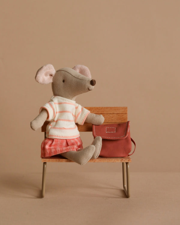 The Maileg Big Sister With Backpack - Coral sits on a small wooden bench, wearing a striped shirt and red shorts. A school backpack placed beside her adds to her charm against the neutral beige background.