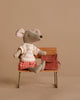 The Maileg Big Sister With Backpack - Coral sits on a small wooden bench, wearing a striped shirt and red shorts. A school backpack placed beside her adds to her charm against the neutral beige background.