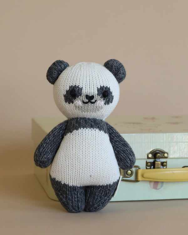 A Cuddle + Kind Baby Panda, crafted from Peruvian cotton yarn, with black and white stripes, sits in front of a pastel background, perched on a stack of light-colored books.