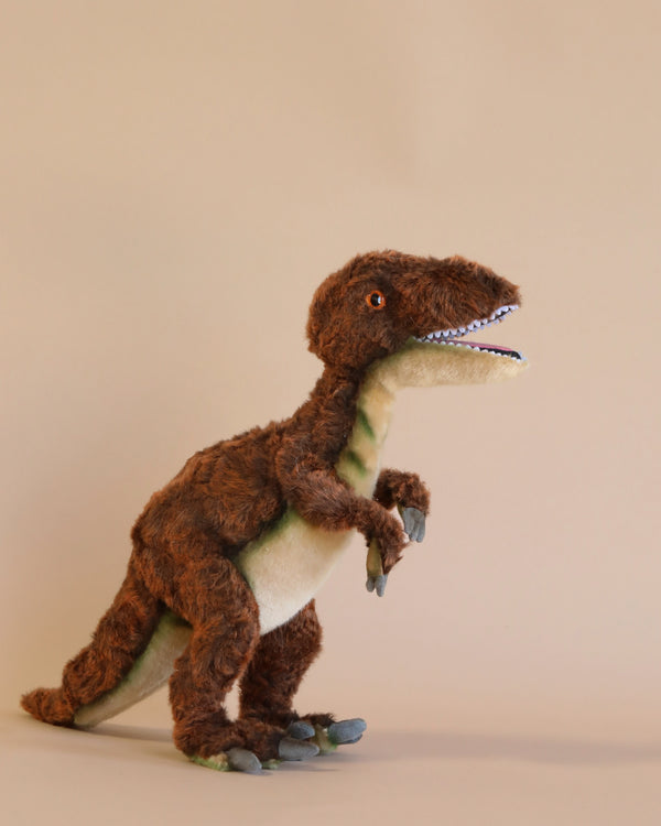 A Velociraptor Dinosaur Stuffed Animal crafted with high-quality materials, featuring brown and green fur, standing upright against a plain beige background. Its mouth is slightly open, showing white teeth.