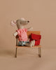 The Maileg Big Sister with Backpack - Red, a petite stuffed mouse in a pink checked dress, sits on a mini wooden bench with her red school backpack nearby against a neutral beige background.