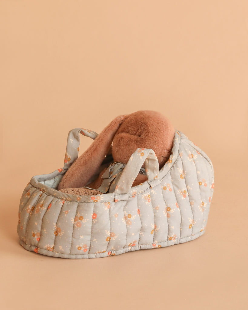 The Maileg Sleeping Bunny in Carry Cot features a soft brown plush bunny with long floppy ears, nestled in a small, padded carry cot decorated with a light blue floral pattern on a beige background.