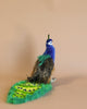 A colorful, whimsical Peacock Stuffed Animal with a vibrant blue and green body and an embellished tail featuring eye-like patterns, positioned against a soft peach background.