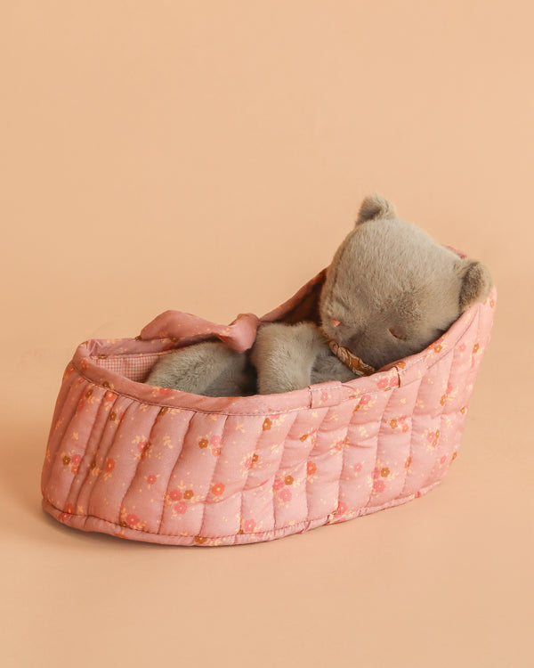A gray Maileg Sleeping Kitten lies on its side in a pink, quilted, floral-patterned carry cot against a peach-colored background.