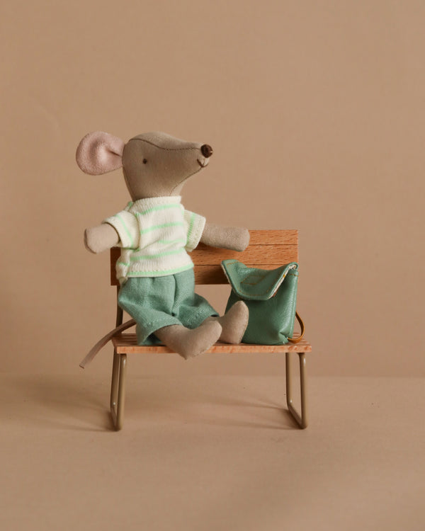 The Maileg Big Brother With Backpack - Mint sits in a striped shirt and green pants on a small wooden bench with metal legs. A matching mint backpack rests beside him, set against a beige background.