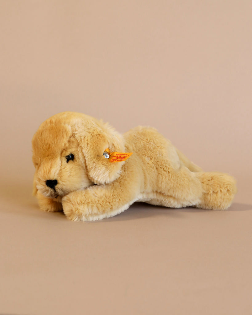 A Steiff Golden Retriever Dog Stuffed Animal, identifiable by the Button in Ear trademark, is lying on its side against a plain beige background. The stuffed animal has soft, realistic fur texture and blue eyes.