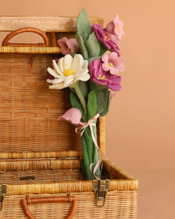 The Handcrafted Felt Flower Bouquet, featuring white, yellow, and pink felt blossoms with green leaves and a ribbon tie, is displayed leaning against an open wicker basket on a beige background.
