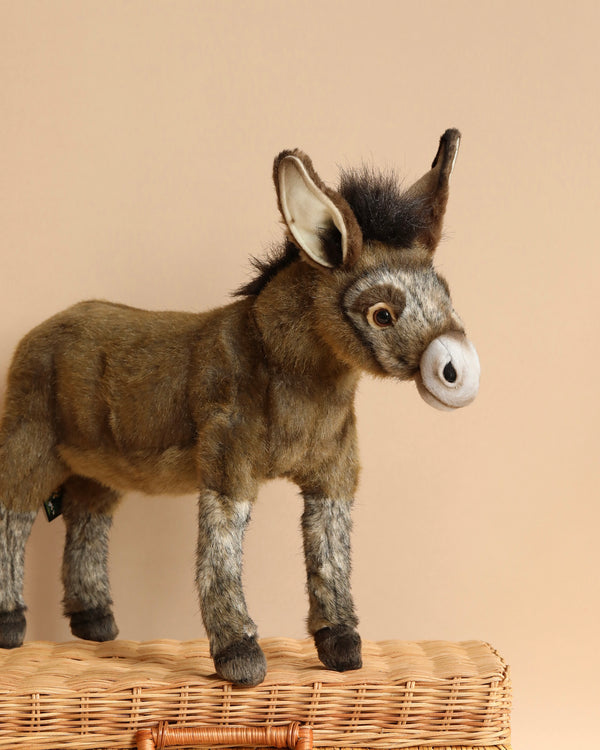 The Donkey Stuffed Animal, characterized by its unique personality and lifelike features, stands on a wicker basket against a beige background. This hand-sewn plush donkey displays brown fur, a white and gray snout, large ears with white insides, and a dark mane on its head.