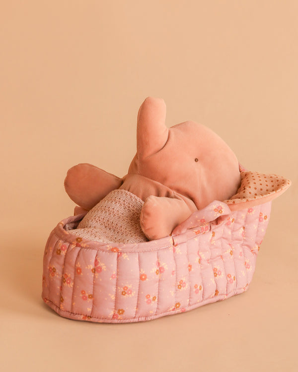 The Maileg Elephant in Carry Cot features a soft pink elephant nestled in a quilted Vintage Rose carry cot with dotted beige lining, all set against a peach background.