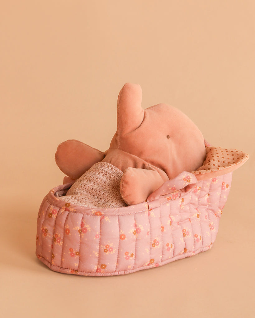 The Maileg Elephant in Carry Cot features a soft pink elephant nestled in a quilted Vintage Rose carry cot with dotted beige lining, all set against a peach background.