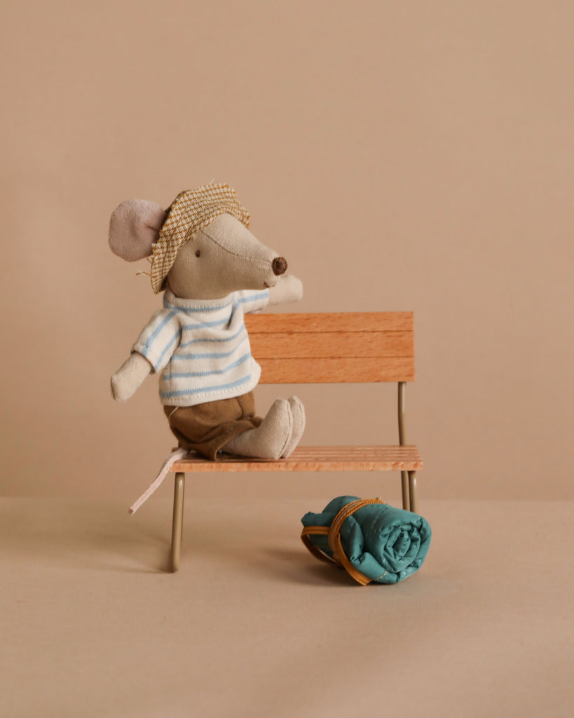 A Maileg Hiker Mouse With Blue Striped Shirt - Big Brother sits on a bench, wearing a striped shirt and hat. A rolled-up blue fabric tied with a string is nearby, possibly as hiking gear. The background is neutral beige.