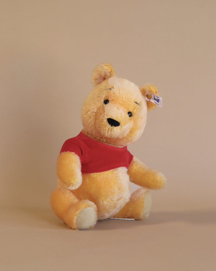 A Steiff Disney's Winnie the Pooh Open Edition Collectible, 10 Inches resembling Winnie the Pooh with fluffy yellow fur, wearing a red t-shirt, sitting against a plain beige background. The bear has a visible tag on its ear.