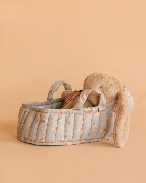 The Maileg Plush Bunny in Carry Cot features a dusty brown bunny with floppy ears resting in a light blue, floral-patterned cot with handles, set on a soft beige background.