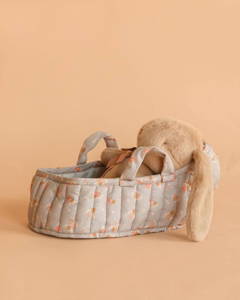 The Maileg Plush Bunny in Carry Cot features a dusty brown bunny with floppy ears resting in a light blue, floral-patterned cot with handles, set on a soft beige background.