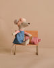 The Maileg Hiker Mouse With Blush Sleeping Bag - Big Sister, from the hiker collection, features a headscarf, pink shirt, and blue dress. It sits on a wooden bench with a pink bag with gold trimming in its lap, against a plain beige background.