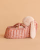 A Maileg Plush Bunny in Carry Cot with long floppy ears rests in a small, pink, quilted floral bed against a plain peach background.