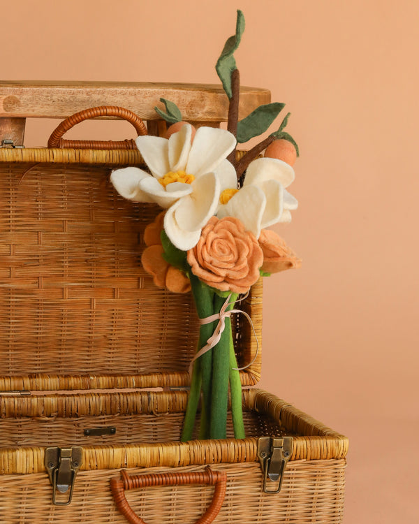 The Handcrafted Felt Flower Bouquet with cream and peach wool-felt flowers and green stems, tied with a ribbon, is displayed in front of an open wicker picnic basket against a peach background.