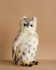 The Snowy Owl Stuffed Animal features hand sewn white and gray fur with black spots, round yellow eyes, and a small beak. It sits upright against a plain beige background, capturing the lifelike detail typical of HANSA creations.