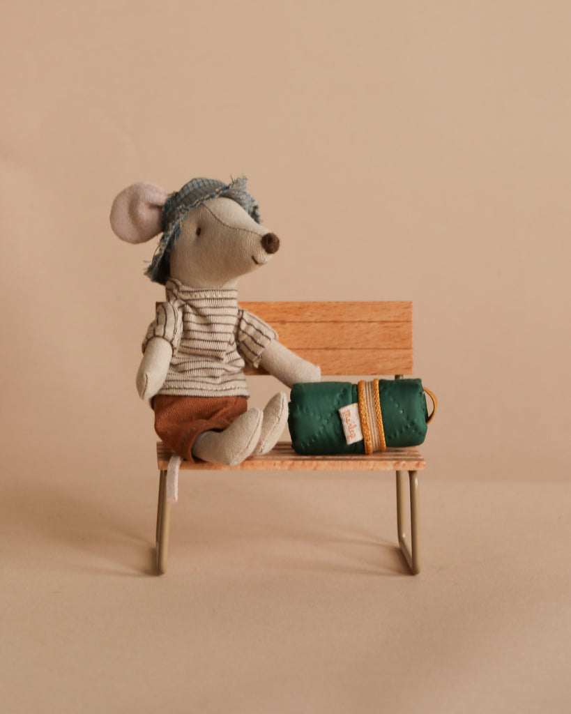 A Maileg Hiker Mouse from the Big Brother collection, wearing a thin striped shirt and brown shorts with magnets in its hands, sits on a wooden bench beside a green bag with a yellow strap against a plain beige background.