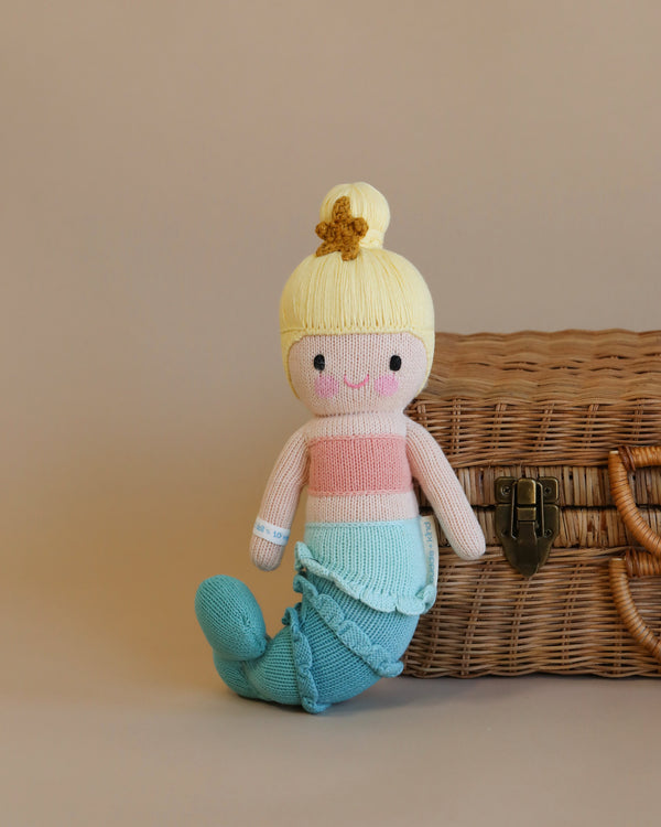 A Cuddle + Kind Skye the Mermaid doll with a blonde bun, pink top, and blue tail sits in front of a wicker basket on a beige background.