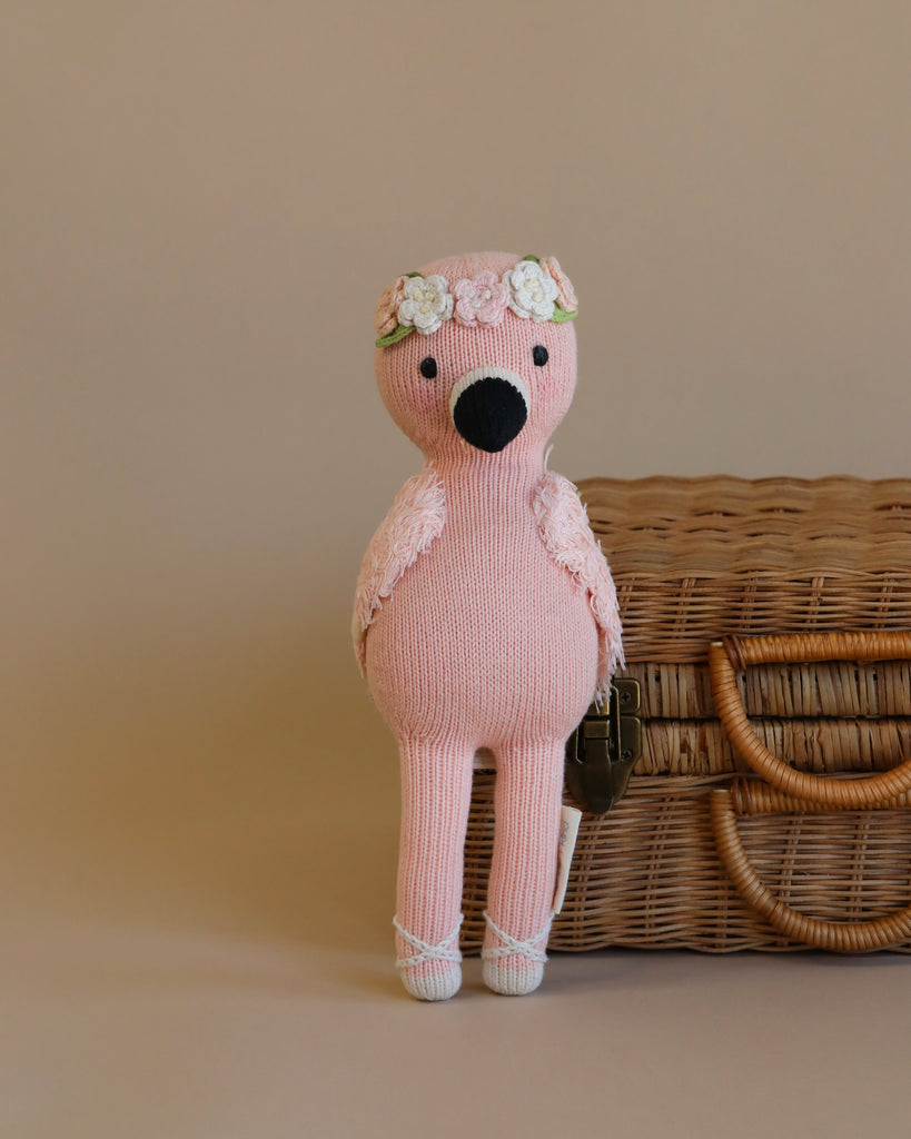 A hand knit pink Cuddle + Kind Penelope the Flamingo with a floral headband sits in front of a wicker basket on a beige background.
