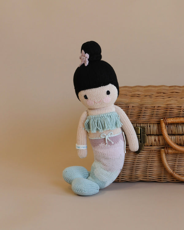 Cuddle + Kind Luna The Mermaid with black hair styled in a bun, wearing a pastel blue and white outfit, sits beside a wicker basket against a neutral background.