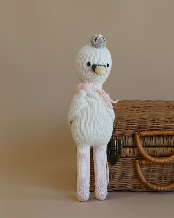 A Cuddle + Kind Harlow The Swan toy with a pink scarf and a gray hat, filled with hypoallergenic polyfill, standing next to a wicker basket on a beige background.