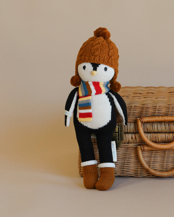 A Cuddle + Kind Everest The Penguin wearing a striped scarf and a pom-pom hat, leaning against a wicker basket on a plain background, crafted using hand knit cotton.