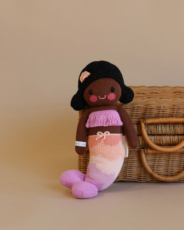 A Cuddle + Kind Maya the Mermaid doll with dark brown skin and black hair sits beside a wicker basket. The doll wears a pink and white striped dress and a purple scarf, with a small flower accessory in her hair.