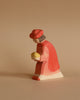 The Ostheimer Red King is a small, wooden figurine of a person wearing a red hat and red coat, and holding a yellow object. Handcrafted from sustainably sourced materials, this simple, minimalist toy is set against a solid beige background.