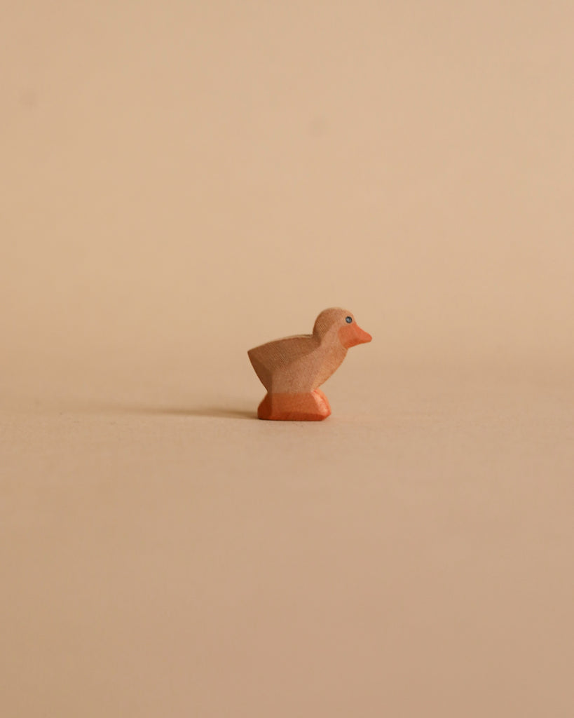 A small, brown wooden chick figurine handcrafted in the style of Ostheimer toys is placed on a neutral beige surface with a matching beige background. Minimalistic in design, the chick is positioned slightly to the right, perfect for imaginative play.