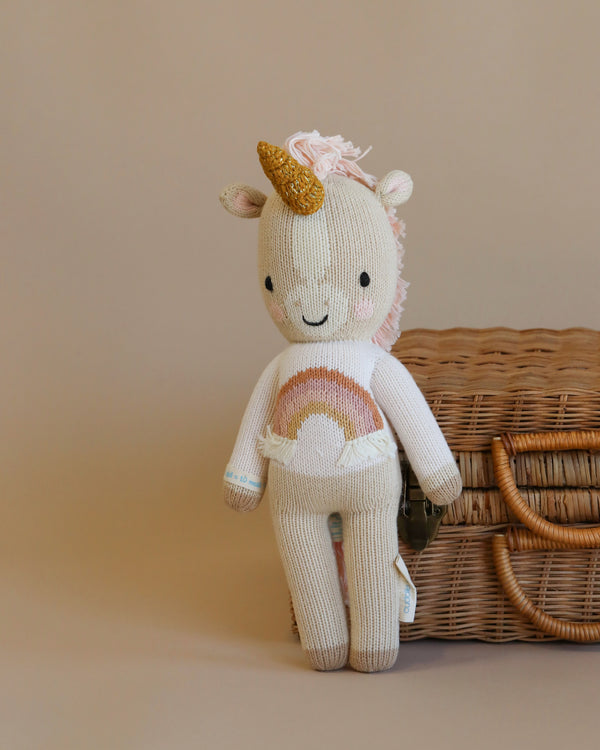 A Cuddle + Kind Zara the Unicorn toy with a golden horn and pastel rainbow details, standing in front of a wicker basket on a beige background.