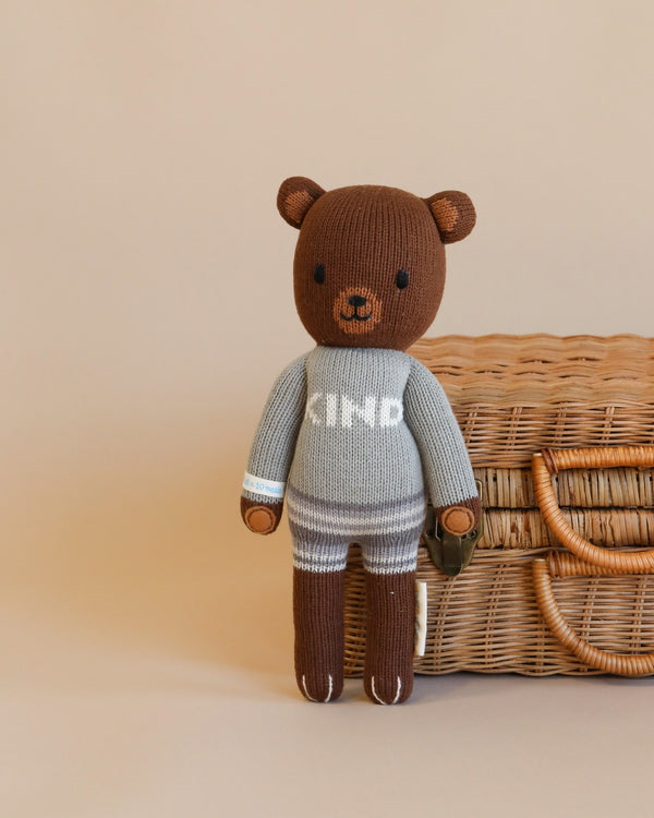 A Cuddle + Kind Oliver the Bear wearing a grey and white sweater with "ted" on it, standing in front of a wicker basket on a beige background.