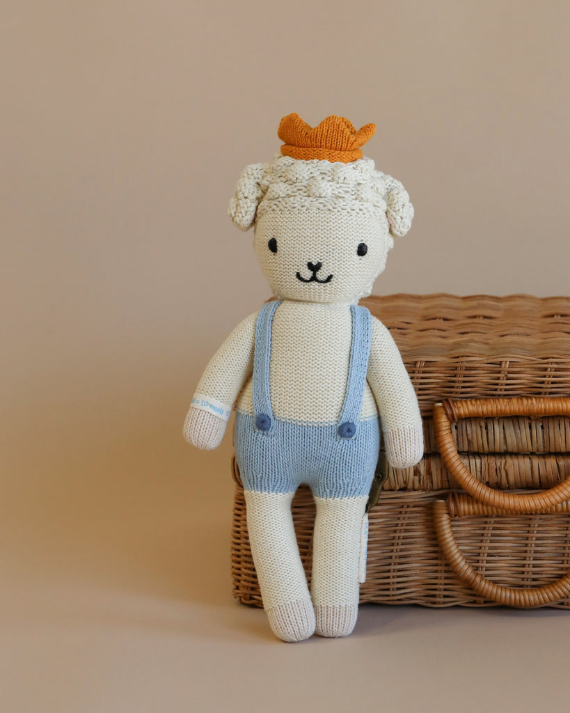 A Cuddle + Kind Lamb Stuffed Animal wearing a blue and white outfit with an orange hat, standing in front of a wicker basket on a beige background, stuffed with hypoallergenic polyfill.