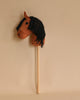 A Hobby Horse brown fabric hobby horse with black yarn mane and black eyes is mounted on a wooden stick against a plain beige background. Made from organic cotton, this delightful toy resembles a classic children's riding stick horse.