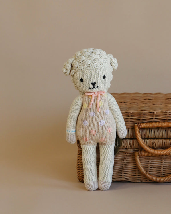 Cuddle + Kind Lucy the Lamb, a hand-knit doll with a cream body, pink and purple polka dots, and a pink bow, is displayed in front of a wicker basket on a beige background.