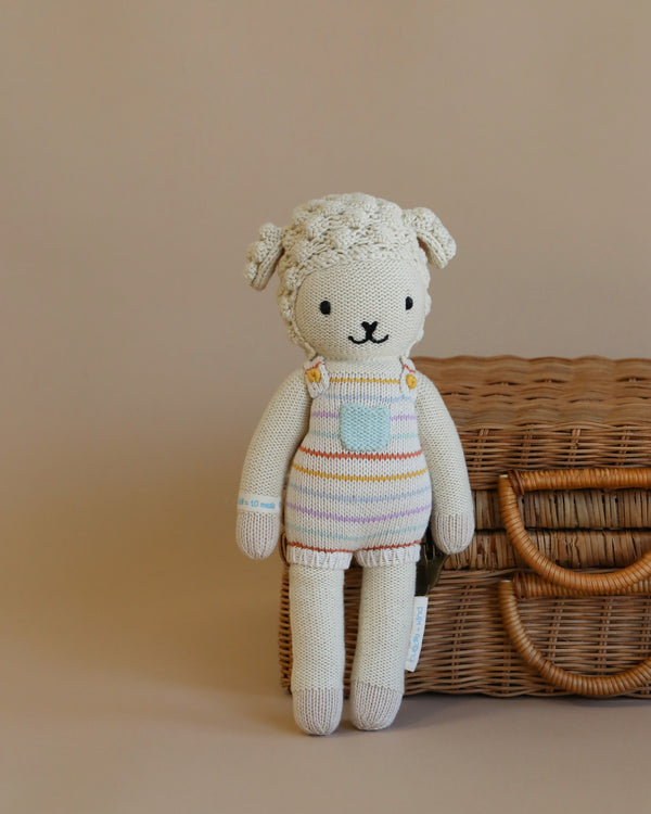Cuddle + Kind Avery the Lamb is a hand-knit, cream-colored plush doll with striped overalls, sitting by a wicker basket on a beige background—crafted using sustainable, fair trade practices.